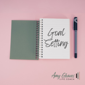 Top-down view of a spiral-bound notebook with "Goal Setting" handwritten on the first page, placed on a light pink surface next to a pen