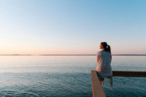 Woman sitting on a wooden dock looking out at a calm body of water during sunset.
