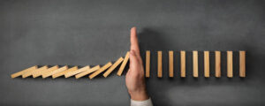 Hand stopping dominoes, it illustrates proactive risk management and intervention to prevent an issue.
